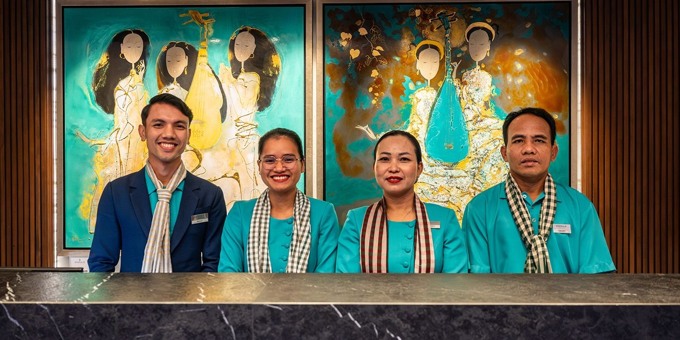 Four Emerald Cruises reception staff in teal outfits and scarves pose at the river cruise reception desk, framed by colorful abstract paintings of musicians in the ship’s lobby.