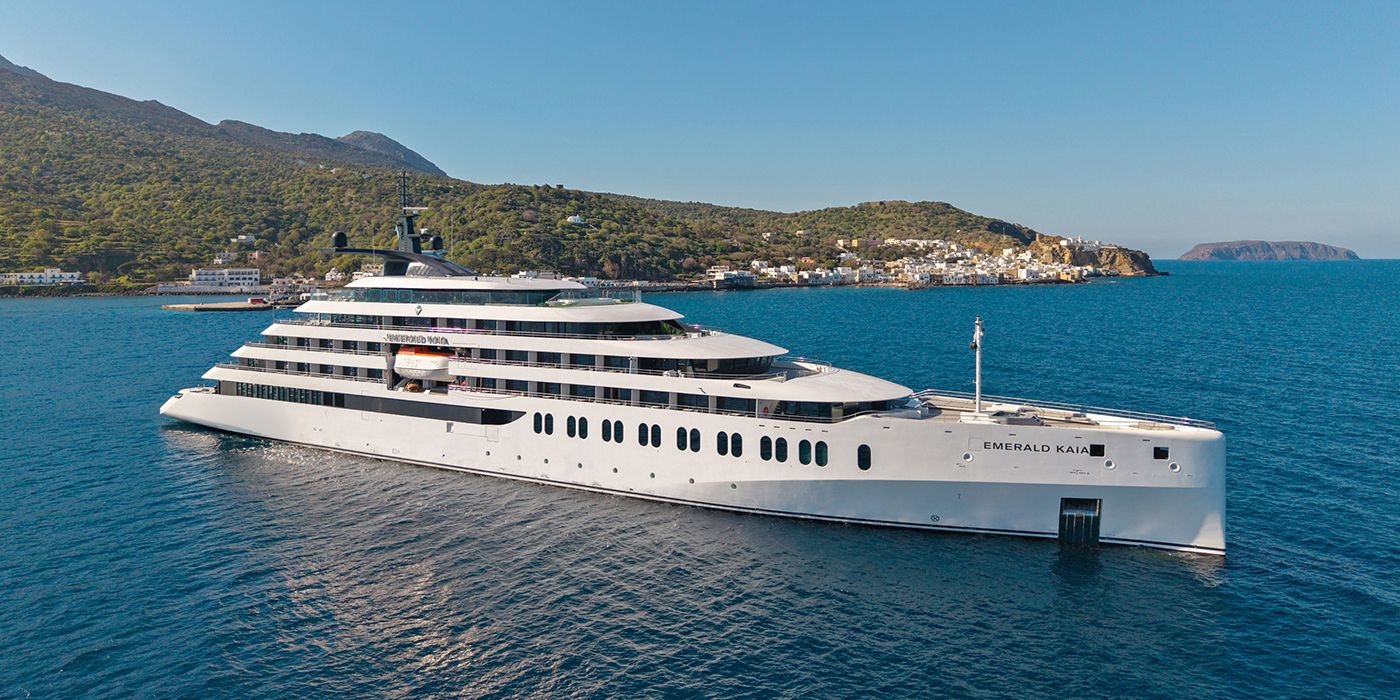 Emerald Kaia superyacht anchored off a green, hilly coastline, its sleek white profile reflected in calm blue water with a coastal village in the background.